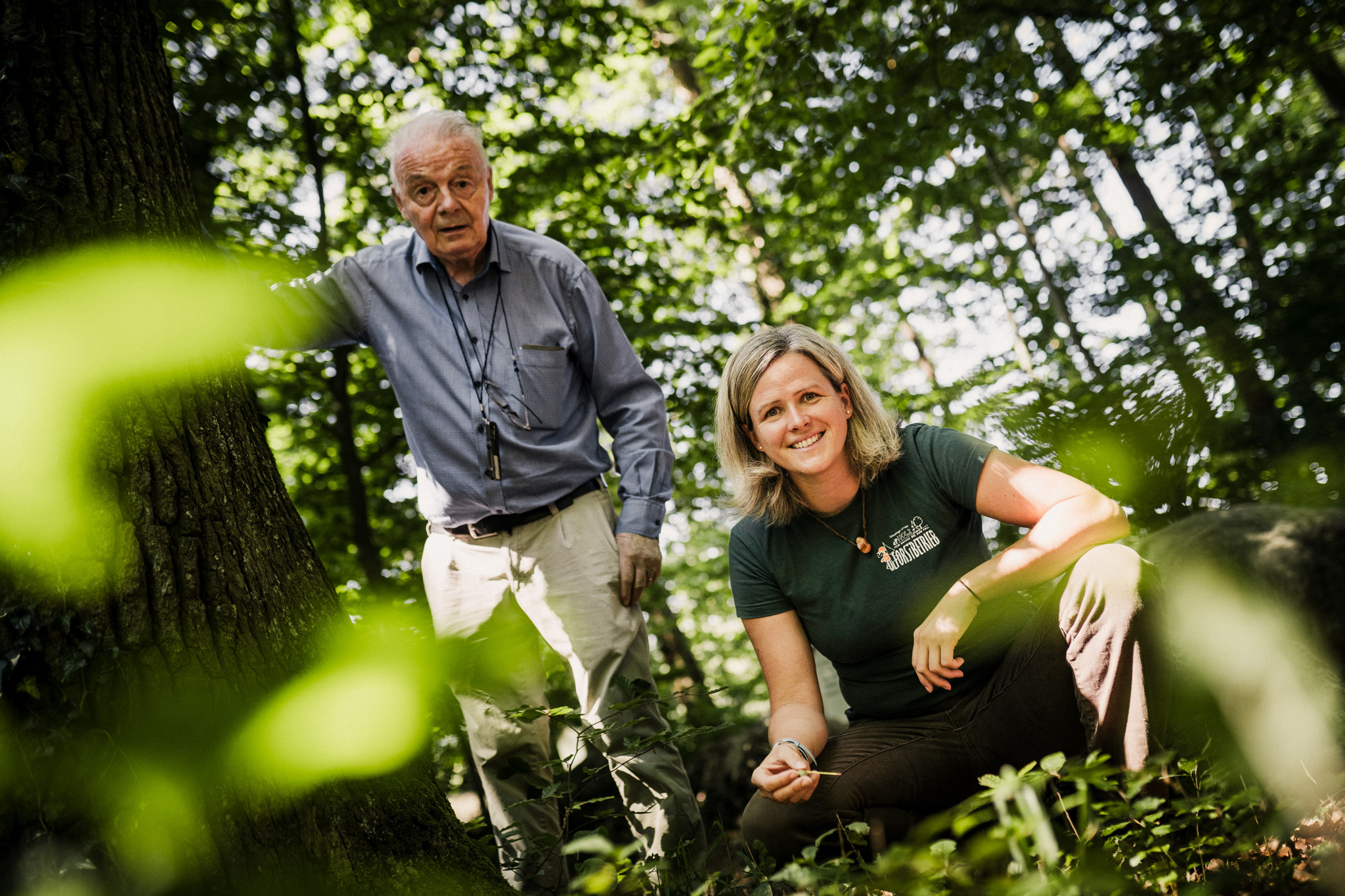 Ein Senior in einem blauen Hemd steht neben einem Baum, während eine Frau in grüner Kleidung lächelnd in die Kamera blickt. Beide befinden sich in einer natürlichen Waldumgebung.